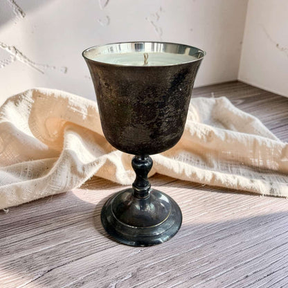 A dark, textured metal candle holder with a single wick, filled with white wax, set on a rustic wooden surface with a draped cream linen cloth in the background.