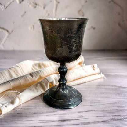 A dark, rustic pewter goblet with a distressed finish, styled on a light gray wooden surface next to a cream-colored linen cloth.