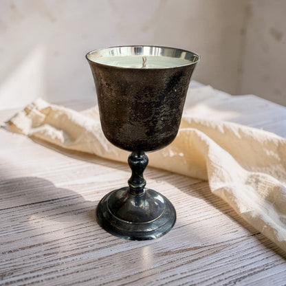 A candle in a dark, textured metal goblet-style holder with a silver rim, resting on a rustic white wooden surface next to a cream-colored linen cloth.