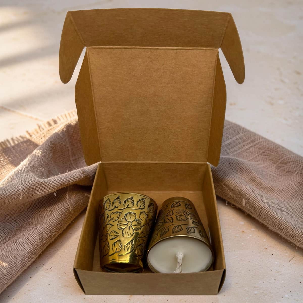 An open kraft paper gift box containing two small decorative candles: one in a floral-patterned brass holder and one white candle in a matching brass holder. The box is styled on a light surface with a textured beige cloth behind it.