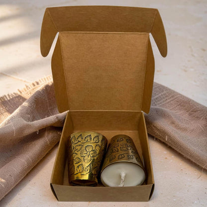 An open kraft paper gift box containing two small decorative candles: one in a floral-patterned brass holder and one white candle in a matching brass holder. The box is styled on a light surface with a textured beige cloth behind it.