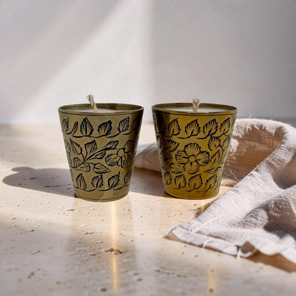 Two small metal candle holders with a gold finish and black floral line patterns, shown on a light stone surface with a folded beige cloth beside them.