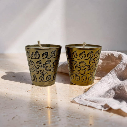 Two small metal candle holders with a gold finish and black floral line patterns, shown on a light stone surface with a folded beige cloth beside them.