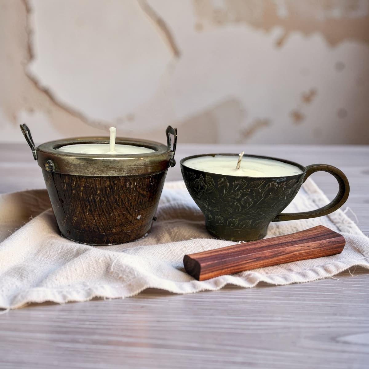 Two rustic metal candle vessels on a cream cloth over a wood surface, with a wooden block in front. One vessel is a small dark bronze pot with side handles and a single wick, and the other is a dark metal cup with a handle and a single wick.