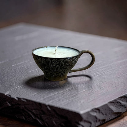A small ceramic candle in a cup-shaped holder with embossed floral detailing, filled with white wax and set on a dark slate board.