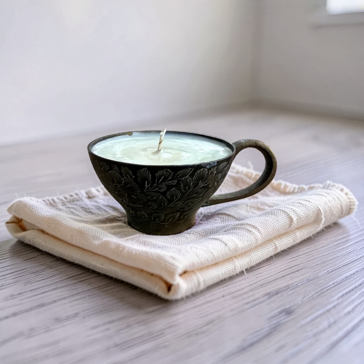 A dark brown carved ceramic candle holder shaped like a teacup, filled with pale green wax and resting on a folded cream-colored textured cloth over a gray wood surface.