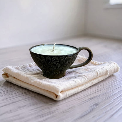 A dark brown carved ceramic candle holder shaped like a teacup, filled with pale green wax and resting on a folded cream-colored textured cloth over a gray wood surface.