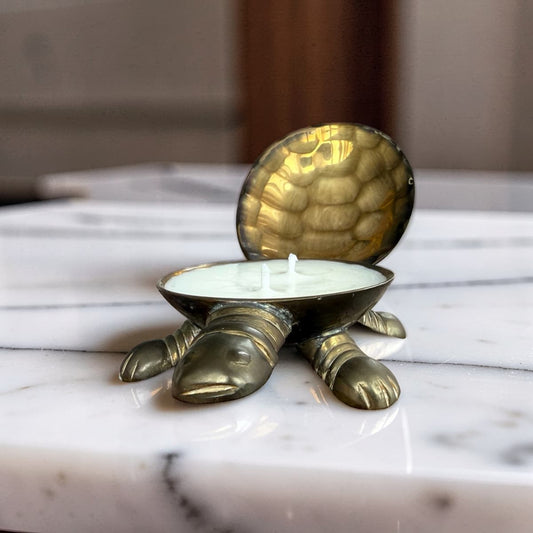 A brass turtle-shaped candle holder with an open shell lid, holding a white wax candle, set on a white marble surface.