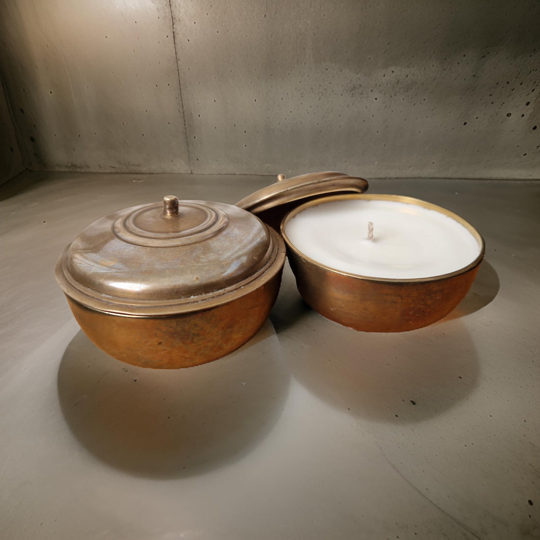 Two bronze containers, one with a lid and the other with a candle inside, on a concrete floor.