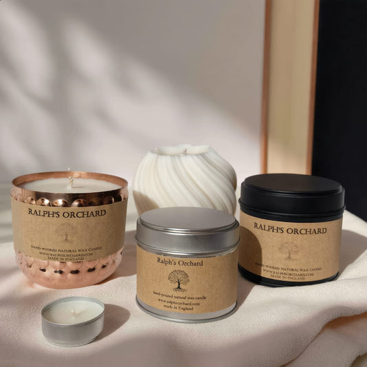 Three Ralph's Orchard unscented candles in different containers (a hammered copper vessel, a white ribbed ceramic vessel, and two silver metal tins with kraft paper labels) arranged on a draped cream fabric with soft shadows.