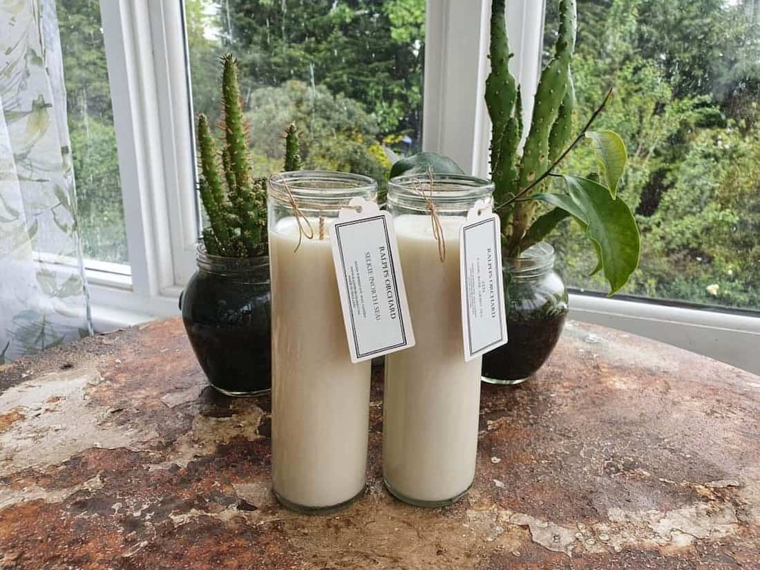 Two candles in glass jars with tags on a rustic surface with plants in the background.