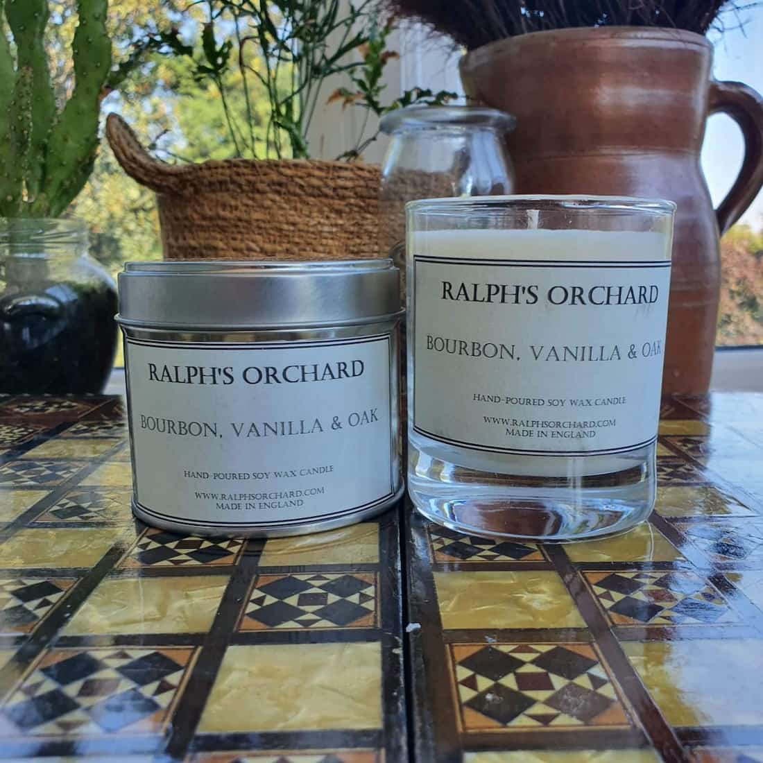 Two candles labeled 'Ralph's Orchard' on a patterned surface with plants in the background.