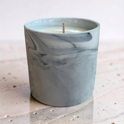 A gray and white marble-patterned glass candle jar with a single wick, set on a light stone surface against a soft neutral background.