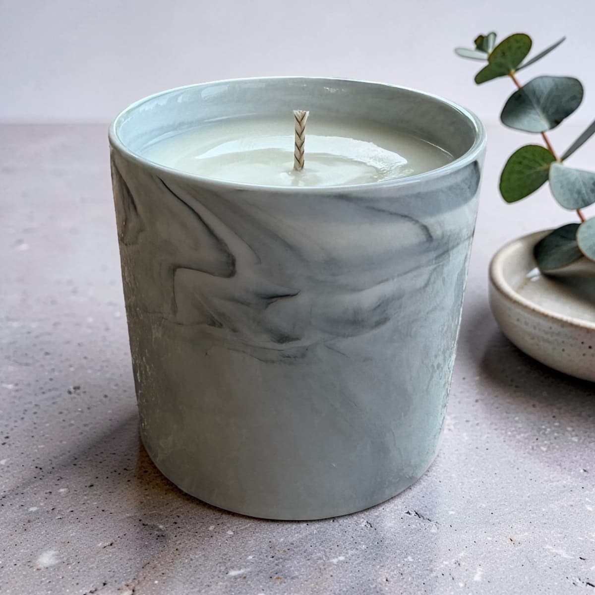 A marble-patterned ceramic candle jar with a light sage green and gray swirl design, filled with white wax and featuring a single wick, set on a textured gray surface next to a small eucalyptus sprig.