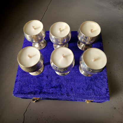 Six white candles in glass holders on a blue fabric with a metal clasp.