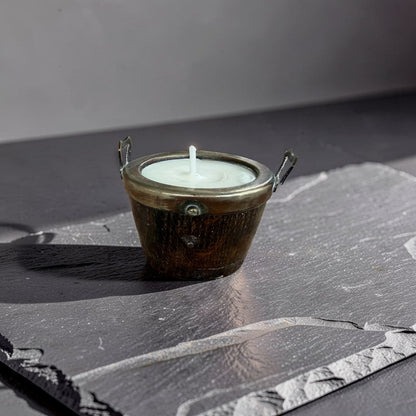 A small bronze metal candle holder with side handles, holding a white candle, placed on a dark slate surface against a gray background.