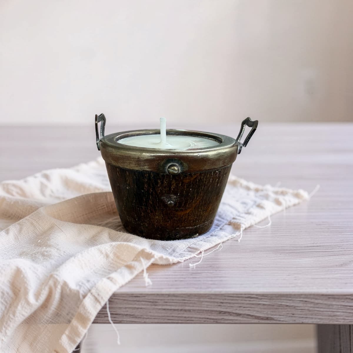 A rustic dark metal candle holder with two side handles and a single white wick, filled with white wax, resting on a light beige linen cloth atop a light wood table.