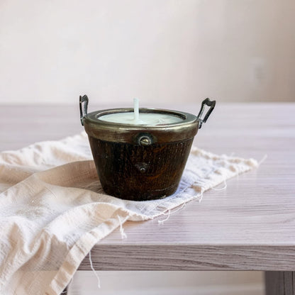 A rustic dark metal candle holder with two side handles and a single white wick, filled with white wax, resting on a light beige linen cloth atop a light wood table.