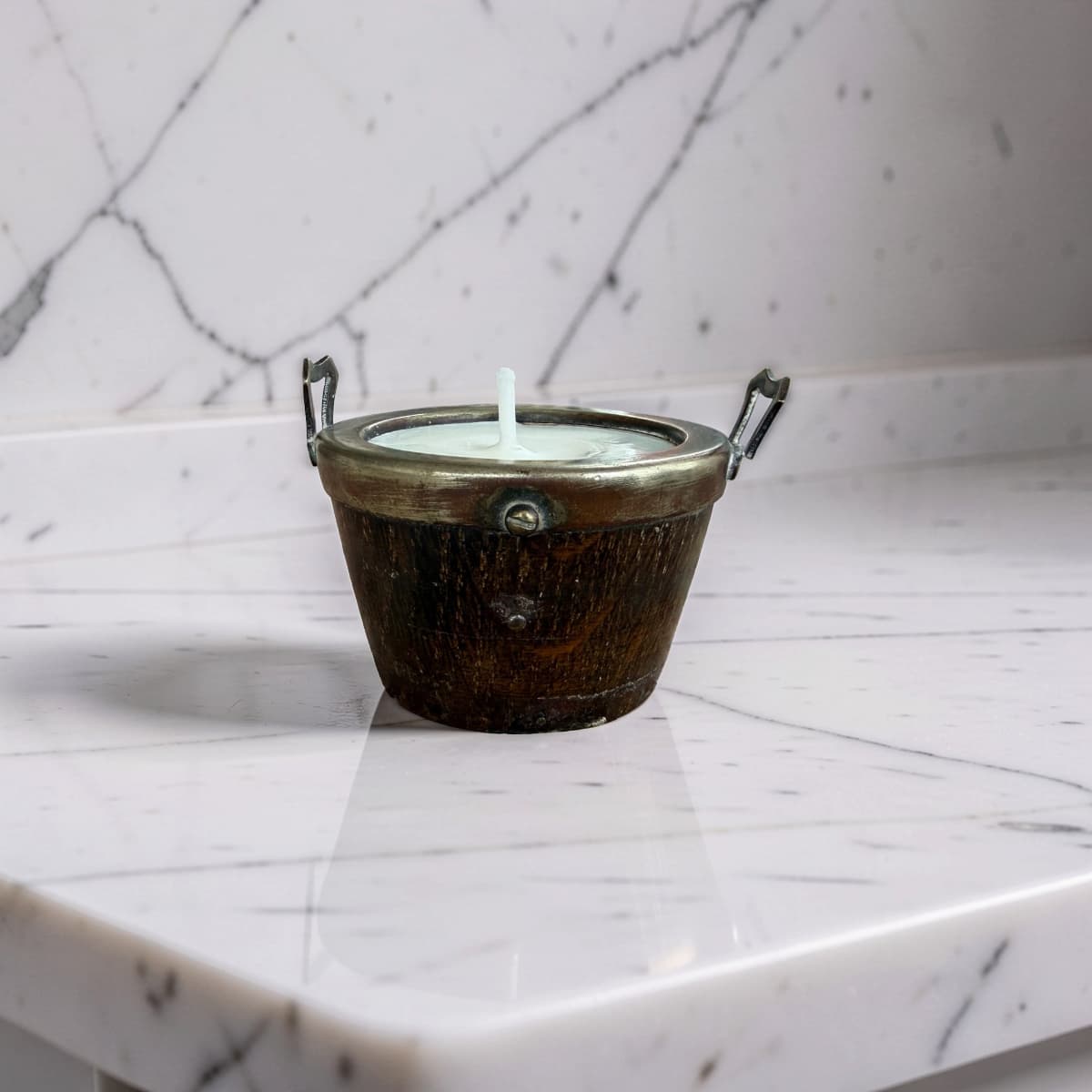 A small metal candle holder with a white wax candle, set on a white marble surface against a matching marble background.